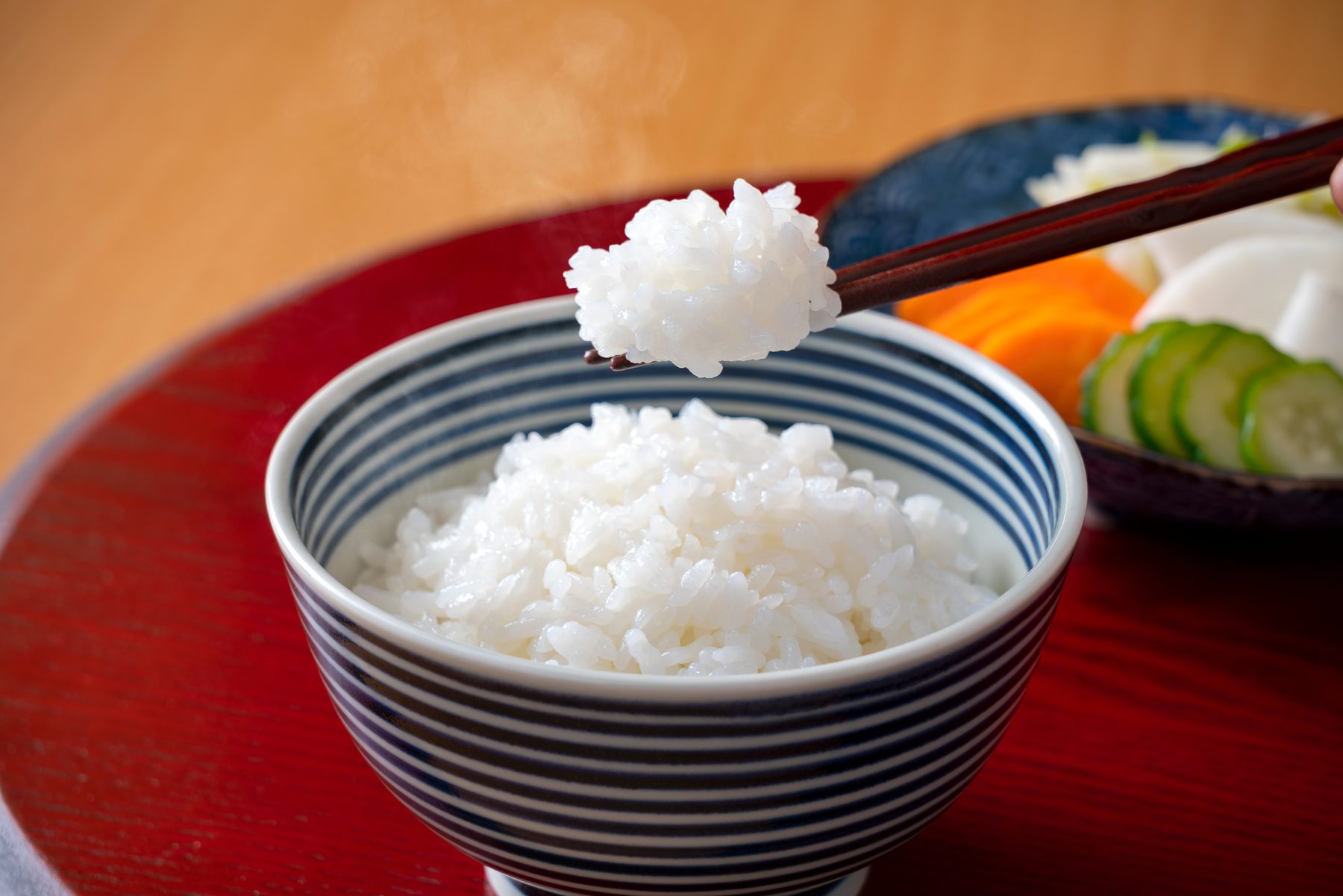 A blue and white striped rice bowl full of white rice, with chopsticks lifting a bite, on a red lacquer tray