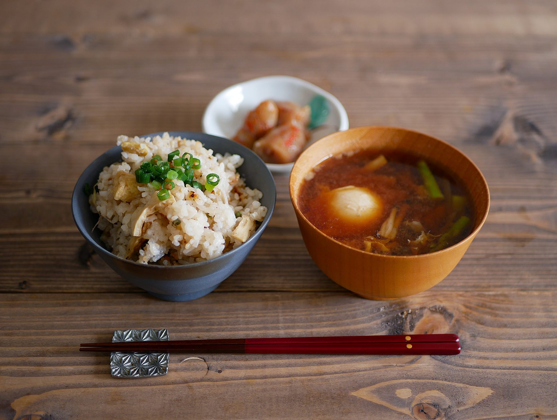 A Japanese meal on a wooden table: rice in a dark bowl on the left, miso soup in a wooden bowl on the right, a small dish of pickles behind, and chopsticks on a rest in front