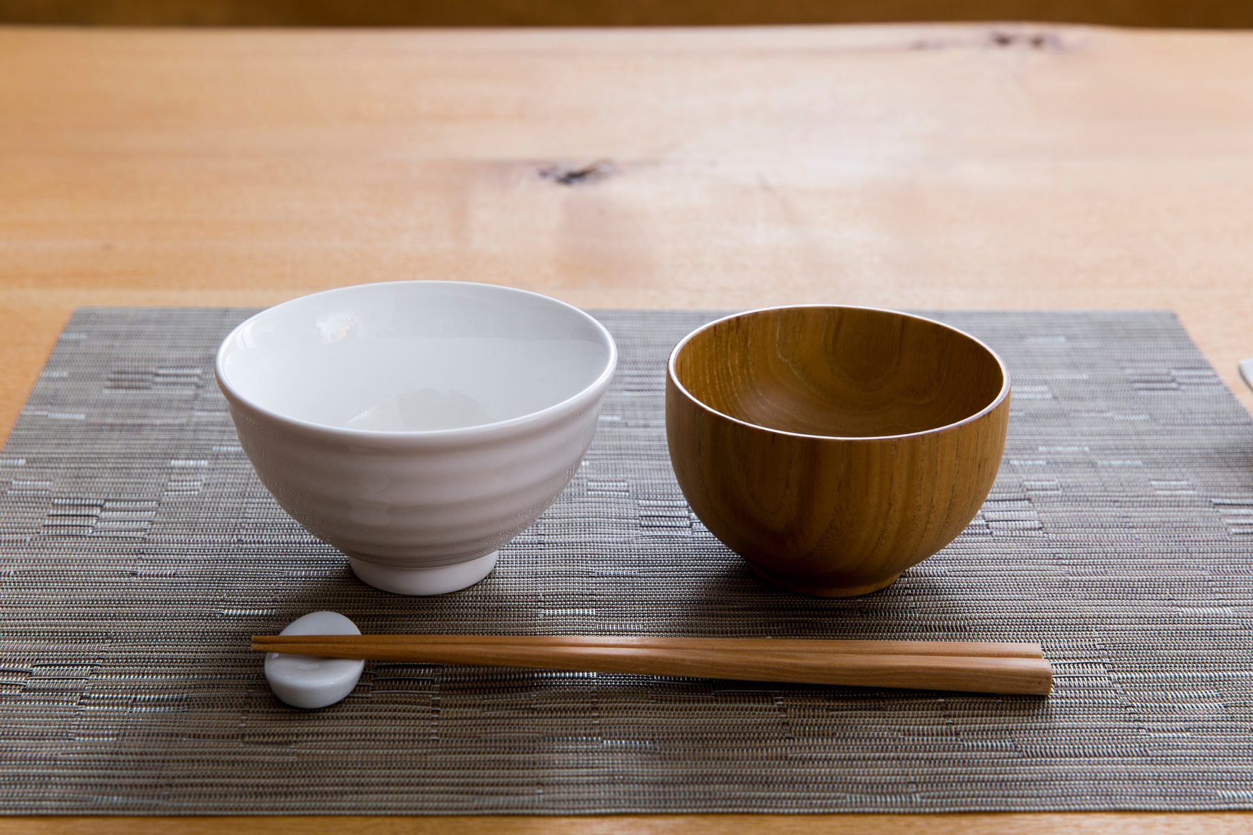 A white ceramic rice bowl and a wooden soup bowl side by side on a woven mat, with chopsticks on a white rest in front
