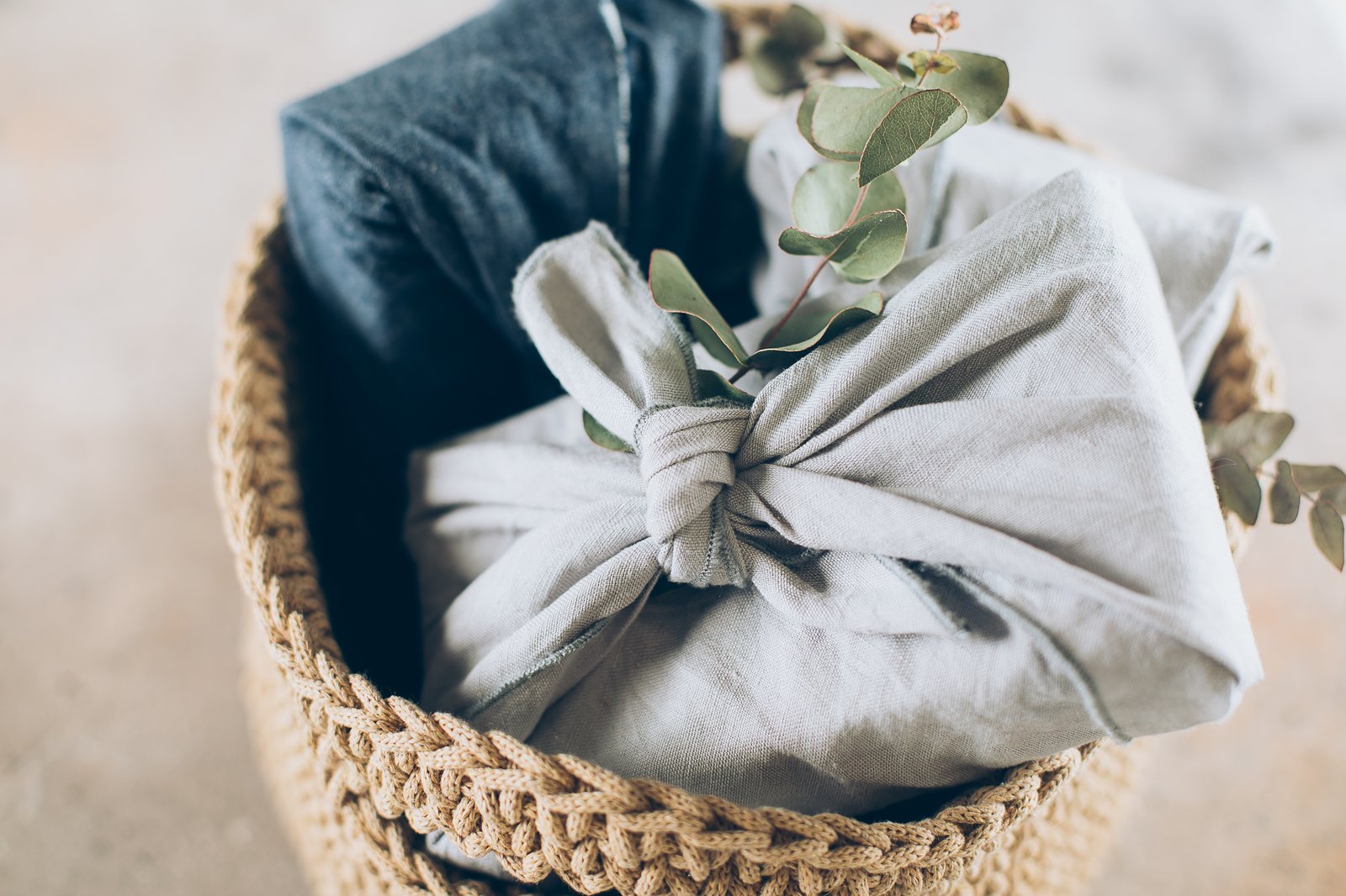 Folded furoshiki cloths in blue and gray tones, resting in a woven basket with a sprig of eucalyptus