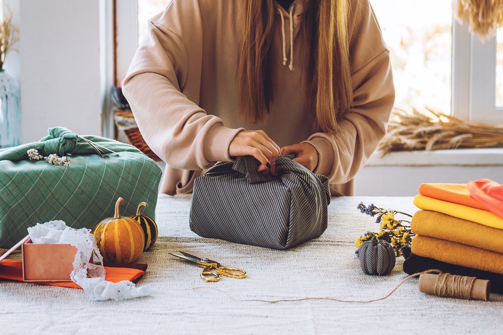 Hands tying a furoshiki cloth around a box, with dried pumpkins and fabric on the table
