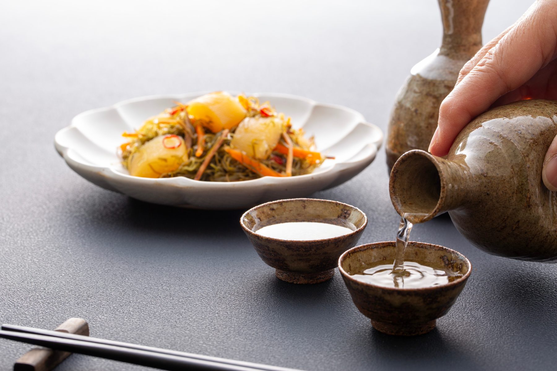 A hand pouring sake from a ceramic tokkuri into small cups next to a plate of simple stewed vegetables, with chopsticks on a dark table