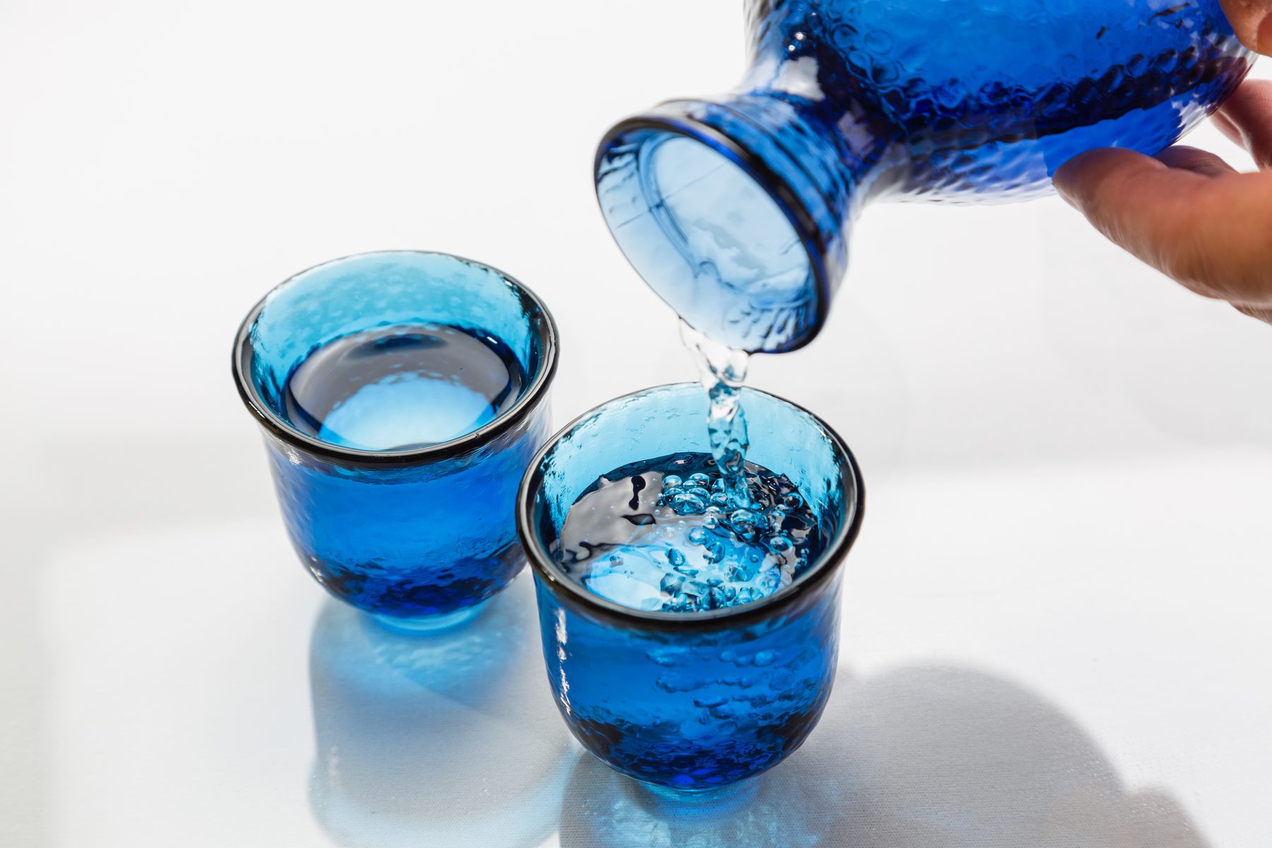Cold sake being poured from a blue glass carafe into two blue glass cups
