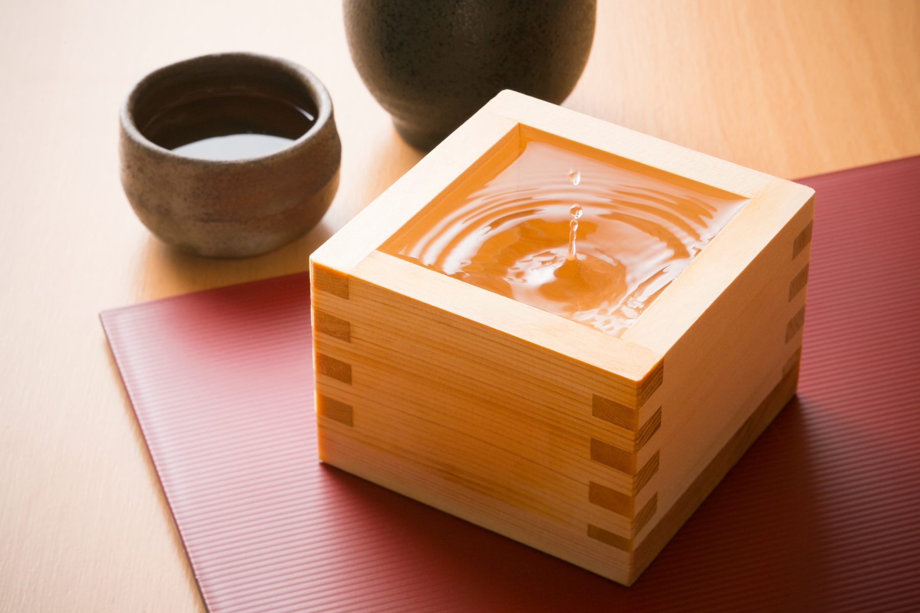 A square wooden masu filled to the brim with sake on a red tray, with a ceramic cup and bottle behind it